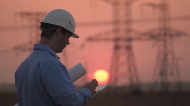 Beyaz kasklı erkek güç mühendisi güç hattını kontrol ediyor, elinde bir tablet tutuyor. Gün batımında yüksek voltajlı elektrik hatları. Dağıtım ve elektrik tedariği. temiz enerji