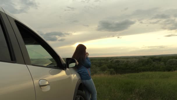 Woman driver traveler resting, drinking morning coffee from a mug in the sun. A tourist girl holds mug of hot tea in her hands and looks at sunset next to car. freedom of travel and tourism by car. — Stock Video