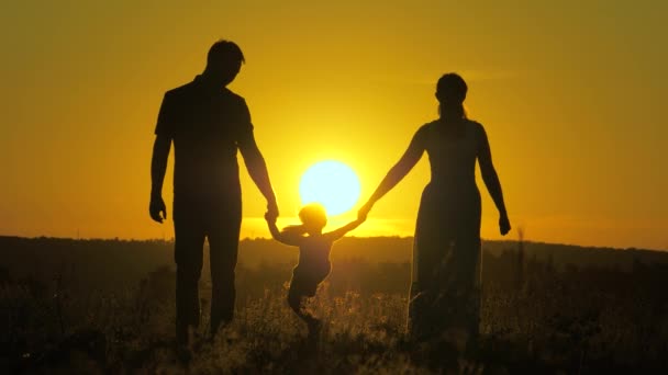 幼い娘が太陽を背景に公園でパパとママの手を握って飛び跳ねる。家族の概念。子供は日没の光の中で父と母と遊ぶ。自然の中で小さな子供と一緒に歩く。幼少期 — ストック動画