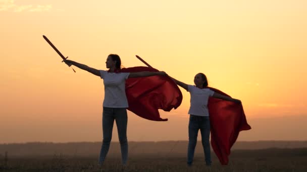 As crianças brincam aos espartanos. meninas de manto vermelho correm com espadas na mão através do campo jogando cavaleiros medievais. As crianças lutam com espada de brinquedo. As crianças brincam aos cavaleiros. meninas saudáveis jogar super heróis — Vídeo de Stock