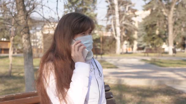 Eine junge Frau in medizinischer Schutzmaske sitzt mit ihrem Smartphone auf einer Straße in Europa. Gesunde Touristinnen auf der Straße tragen Schutzmasken vor Viren. Konzept Gesundheit. — Stockvideo