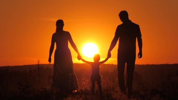 子供は日没の光の中で父と母と遊ぶ。幸せな家族だ。幼い娘が太陽を背景に公園でお父さんとお母さんの手を握ってジャンプします。幼少期 — ストック動画