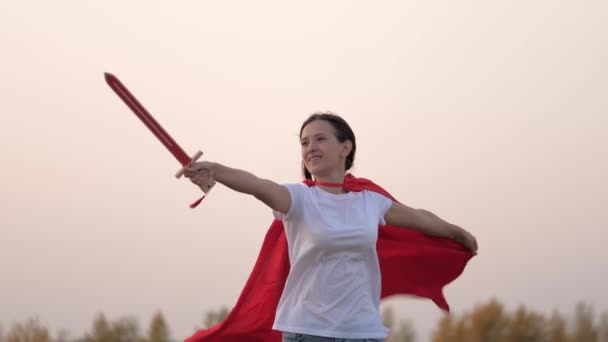 Dětští rytíři. Svobodná dívka v červeném plášti běhá s meči v ruce přes pole a hraje si na středověkého rytíře. Mladá dívka si hraje na superhrdiny. Děti se perou mečem. Šťastné dětství — Stock video