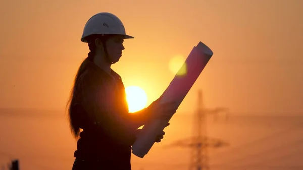 Bir mühendisin silueti yüksek voltajlı bir elektrik santrali inşa projesine bakıyor. Mühendis çizimi okuyor. Mühendis gün batımında elektrik direklerinin yanındaki alanda çalışıyor..