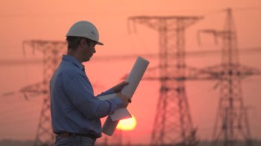Elektrik ve yüksek voltajlı elektrik hatları gün batımında. Beyaz miğferli güç mühendisi güç hattını kontrol ediyor, tablet ve mühendislik diyagramı tutuyor. Dağıtım ve elektrik tedariği. temiz enerji