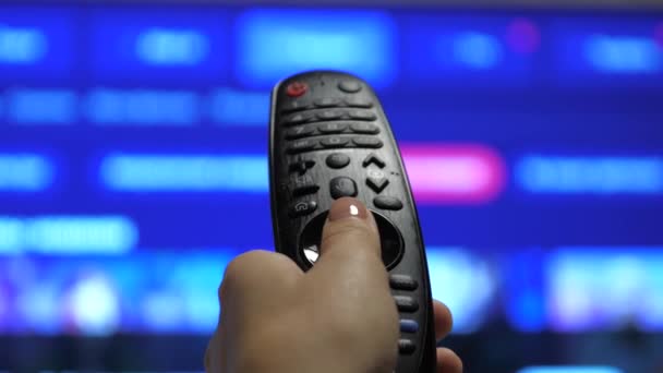 Una persona controlla la TV utilizzando un moderno telecomando. Un uomo guarda una smart TV e usa un telecomando nero. pagine di scorrimento tv sfocate — Video Stock