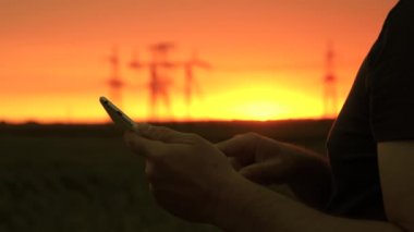 Elinde bir gezegen olan elektrikçi bir elektrik hattında çalışıyor. Yakın plan. Güç mühendisi güç hatlarını kontrol ediyor, elinde bir tablet tutuyor. Gün batımında yüksek voltajlı elektrik hatları. Elektrik tedariği.