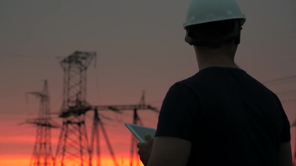 Elektrikçi elektrik hattında çalışıyor. Beyaz kasklı bir enerji mühendisi güç hattını kontrol ediyor, tableti elinde tutuyor. Gün batımında yüksek voltajlı elektrik hatları. Elektrik dağıtımı ve tedariği. — Stok video