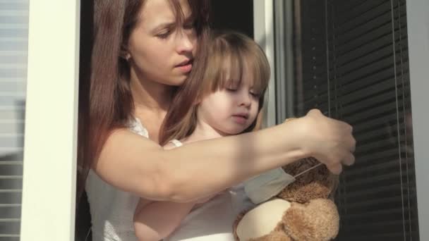 Familjebarn och mor i karantän. Mamma och dotter leker på fönsterbrädan, klär nallebjörn med skyddande mask från virus, tittar ut genom fönstret. leksak med barnkramar. Covid-19 coronavirus — Stockvideo