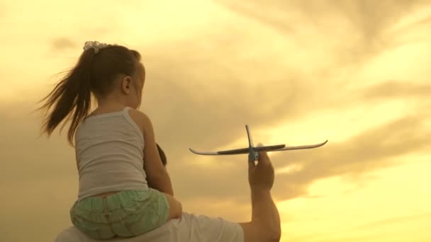 La piccola figlia sulle spalle di papà sta giocando con un aeroplano giocattolo sul campo al sole. bambino e papà giocano un aeroplano giocattolo. il bambino sogna di volare e diventare un pilota e viaggiare. famiglia felice. — Video Stock