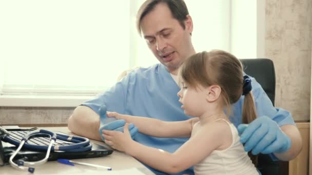 Médico pediatra jugando en su oficina con una niña linda y trabajando en un ordenador portátil. La paciente se sienta en el regazo del pediatra. concepto de examen pediátrico para el tratamiento de niños. — Vídeos de Stock