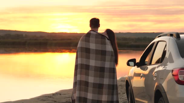 Ευτυχισμένος ερωτευμένοι ταξιδιώτες άντρας και γυναίκα καλύπτονται με κουβέρτα δίπλα στο αυτοκίνητο και να θαυμάσουν όμορφο ηλιοβασίλεμα στην παραλία. τουρίστες δίπλα στο αυτοκίνητο, αγκαλιάζει, θαυμάζοντας την ανατολή του ηλίου ποταμού. Ελεύθεροι ταξιδιώτες. ταξίδι με αυτοκίνητο. — Αρχείο Βίντεο