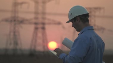 Beyaz kasklı erkek güç mühendisi güç hattını kontrol ediyor, tableti elinde tutuyor. İş adamı çalışıyor. Gün batımında yüksek voltajlı elektrik hatları. Dağıtım ve elektrik tedariği. Temiz enerji