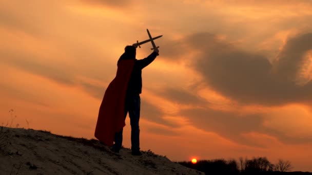 L'uomo libero gioca a fare il supereroe. Un cavaliere maschio libero incrociò le spade nel cielo sopra la sua testa. Superman con la spada in mano e il mantello rosso si erge sulla montagna alla luce del tramonto. Gioco della legione romana. — Video Stock