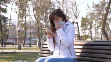 Tıbbi koruyucu maskeli genç bayan gezgin şehir caddesinde akıllı telefonuyla oturuyor. Telefonlu, koruyucu maskeli, virüslü bir turist. Sağlık ve güvenlik kavramı. Coronavirus koruması.