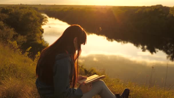 Jovem viajante lê um livro em uma parada. turista menina com um livro sobre a montanha ao sol sobre o rio. Um estudante lê um livro didático no parque ao ar livre em raios de sol. Oração na natureza. — Vídeo de Stock