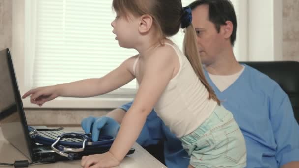 Лікар педіатр грає у своєму офісі з милою маленькою дівчинкою і працює на ноутбуці. Пацієнтка сидить на колінах педіатра. концепція педіатричного огляду для лікування дітей . — стокове відео