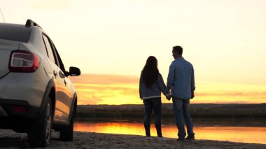 Mutlu aşıklar, gezginler arabanın yanında durur ve sahilde gün batımını seyrederler. Turistler arabayla gezer, kucaklaşır, güneşin doğuşuna ve nehre hayran kalırlar. Bedava gezginler, turistler. Aile seyahati.