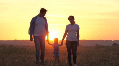 Mutlu aile parkta yürüyor. Güneşin altında el ele tutuşuyorlar. Anne, baba ve çocuk gün batımında mutlu, kız anne ve babasını elleriyle tutuyor. Sağlıklı çocuk atlıyor, ailesiyle oynuyor. Mutlu aile