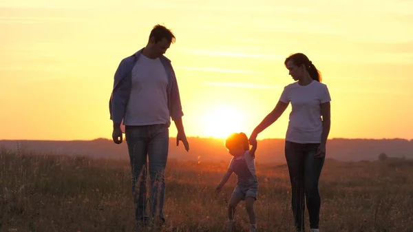 Ευτυχισμένη οικογένεια είναι το περπάτημα στο πάρκο στον ήλιο κρατώντας τα χέρια. Μαμά, ο μπαμπάς και το παιδί είναι ευτυχισμένοι, στο ηλιοβασίλεμα η κόρη κρατάει τους γονείς της από τα χέρια. υγιές παιδί πηδάει, παίζει με τους γονείς του. Ευτυχισμένη οικογένεια — Φωτογραφία Αρχείου