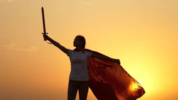 Rapariga joga super-heróis. Criança joga Spartan In. Uma menina livre com um manto vermelho com uma espada na mão interpreta um cavaleiro medieval ao sol. Rapariga luta com uma espada de brinquedo. Conceito de infância feliz. — Fotografia de Stock