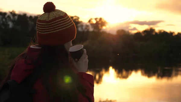 Donna Traveler tiene tazza di metallo con caffè caldo nelle sue mani e guarda il tramonto vicino al fiume. viaggiatore ragazza libera ammirando il paesaggio. Un turista beve il tè da una tazza al sole. viaggi, escursioni — Foto Stock