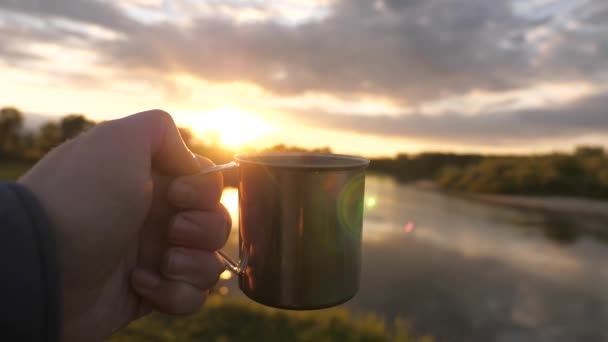 Een toerist drinkt thee uit een mok in de zon. De reiziger hand houdt een metalen mok met hete koffie en kijkt naar de zonsondergang. Een close-up. Avontuur, reizen, toerisme en kamperen. — Stockvideo