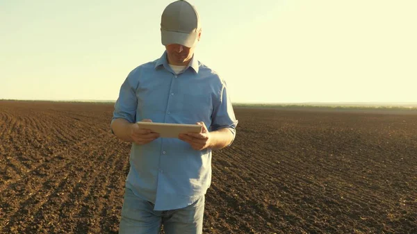A farmer works with a tablet in a plowed field in the sun. agronomist with a tablet studying the harvest of sowing grain in the field. a business person plans his income in this area. grain harvest.