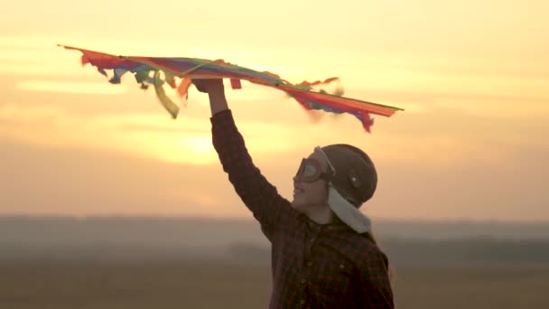 Happy child in a pilots helmet runs with a multi-colored kite in his hands at sunset. girl wants to become pilot. child dreams of freedom, flight and travel. teenager playing on a plane outdoors. — Stock Video