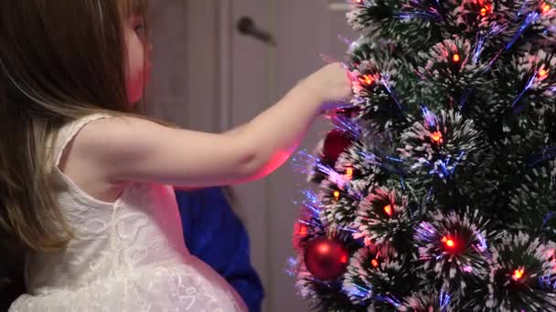 Malá dcera a matka pověsí červenou hračku na vánoční stromek. Šťastnou rodinu, Vánoce. Dítě a máma zdobí vánoční stromeček s vánoční koule. Veselé dítě a maminka si hrají u stromu. — Stock video