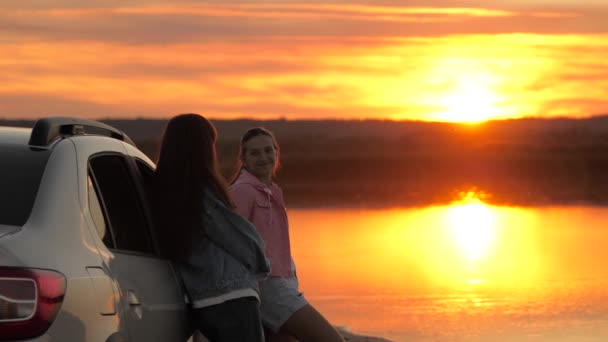 Las madres felices y la hija abrazan, viajan, se paran al lado del coche y admiran la hermosa puesta de sol en la playa. Mujeres turistas libres en coche, admirando el amanecer, río. Viajeros familiares, turistas. Viajes en familia en coche. — Vídeos de Stock