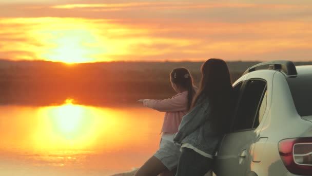 Mutlu anne ve kız kucaklaşır, seyahat eder, arabanın yanında durur ve sahilde gün batımını seyrederler. Özgür kadın turistler arabayla, güneşin doğuşuna, nehre hayran. Aile gezginleri, turistler. Aile seyahati. — Stok video