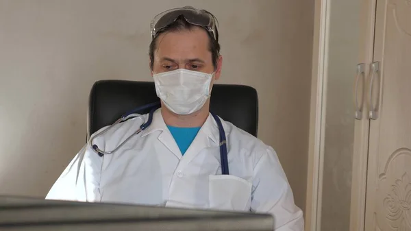 Доктор в лабораторному пальто, окуляри і стетоскоп навколо шиї, працює з ноутбуком за столом. Сімейний лікар працює на комп'ютері в клініці. Робота в медичному кабінеті. Терапевт в лікарні . — стокове фото