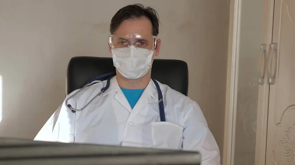 Doctor en bata de laboratorio, gafas y estetoscopio alrededor de su cuello, trabaja con el ordenador portátil en la mesa. El médico de familia trabaja en la computadora de la clínica. Trabaja en un consultorio médico. Terapeuta en el hospital. —  Fotos de Stock