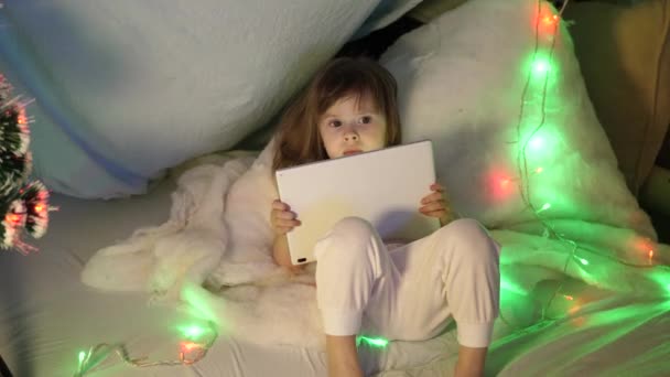 Un niño juega en una tableta en Nochebuena. Linda niña con un gadget en el sofá en la habitación de los niños. Hija mira dibujos animados en el PDA, Niño en la víspera de Año Nuevo en la habitación — Vídeo de stock