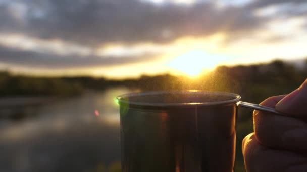 観光客は太陽の下でマグカップからお茶を飲んでいます。旅行者の手は熱いコーヒーと金属マグカップを保持し、日没を見てください。接近中だ。アドベンチャー、旅行、観光、キャンプのコンセプト. — ストック動画
