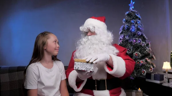 Père Noël est assis sur le canapé avec une petite fille, enfant ouvre cadeau, se réjouit et câlin grand-père magique. Fêtes et célébrations concept. Famille enfants vacances d'hiver. Bonne soirée de Noël. — Photo