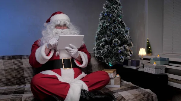 Santa Claus works remotely with a tablet online. Happy elderly man in santa claus costume smiling and looking at modern tablet sitting on sofa near christmas tree in evening in room. New Year Holidays — Stock Photo, Image