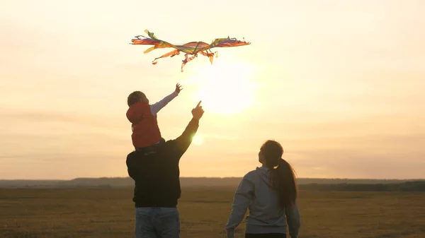 Papa et maman jouent avec leur fille et un cerf-volant dans le parc. Bonne famille. Un enfant sur les épaules de son père attrape joyeusement un cerf-volant avec sa main. Famille lançant des avions en papier coloré dans le ciel — Photo