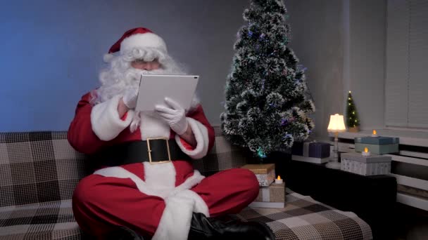 Święty Mikołaj pracuje zdalnie z tabletem online. Szczęśliwy starzec w kostiumie Świętego Mikołaja uśmiechający się i patrzący na nowoczesną tabliczkę siedzącą na kanapie w pobliżu choinki wieczorem w pokoju. Wakacje noworoczne — Wideo stockowe