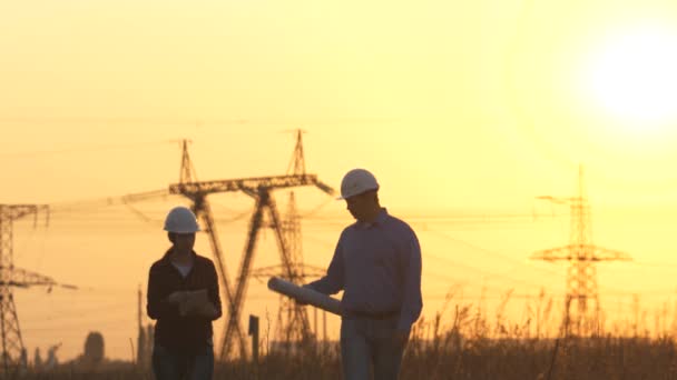 Silhouette af ingeniører ser på opførelsen af højspændingskraft. Teams ingeniør leder diskuterer plan. To ingeniør stående på banen med el-tårne ved solnedgang – Stock-video