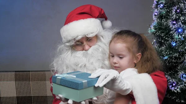 Santa Claus is sitting on couch with a little girl, child opens gift, rejoices and hugs magic grandfather. Holidays and celebrations concept. Family childrens winter holiday. Happy Christmas evening. — Stock Photo, Image