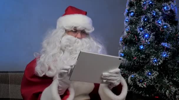 Père Noël fonctionne à distance avec une tablette en ligne. Heureux homme âgé en costume de Père Noël souriant et regardant tablette moderne assis sur le canapé près de l'arbre de Noël dans la soirée dans la chambre. Vacances de Nouvel An — Video