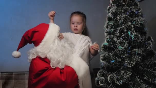 Der Weihnachtsmann sitzt auf der Couch und spielt mit dem kleinen Mädchen. Kind und Nikolaus betrachten und schmücken den Weihnachtsbaum. Feiertage und Feiern. Winterurlaub für Familien mit Kindern. Frohe Weihnachten. — Stockvideo