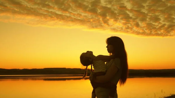 Familia feliz, mamá abrazándose al atardecer con la pequeña hija feliz en la playa. Mamá juega con su hijo junto al agua al atardecer. Concepto familiar feliz. Silueta de madre y bebé saludable bailando. —  Fotos de Stock