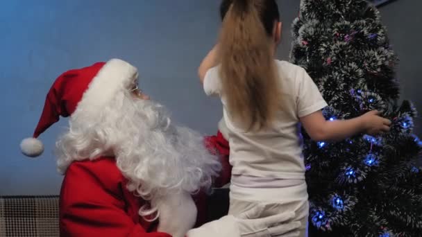 Dítě a Santa Claus se dívají a zdobí vánoční stromeček. Santa Claus sedí na gauči a hraje si s malou holčičkou. Svátky a oslavy. Rodinné děti na zimní dovolenou. Šťastný Štědrý večer.