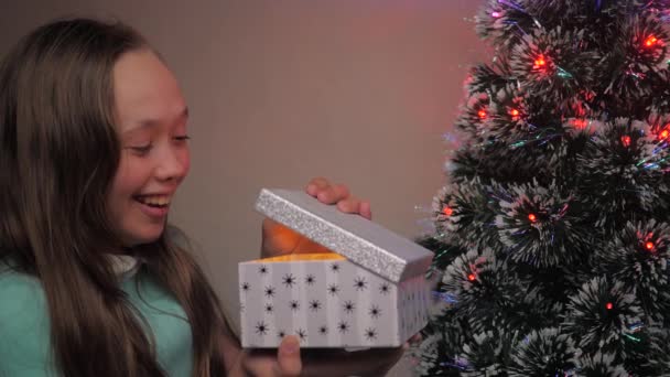 Dziewczynka otwiera prezent od Świętego Mikołaja obok choinki. Dziecko otwiera pudełko prezentów w świetle girlandy, raduje się i uśmiecha. Rodzinne wakacje i uroczystości, zimowy odpoczynek. Wesołych Świąt. — Wideo stockowe