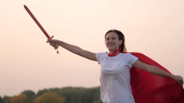 Genç kız süper kahramanları oynuyor. Kız oyuncak kılıçla dövüşüyor. Çocuk Spartan 'ı oynuyor. Elinde kılıç olan kırmızı pelerinli özgür bir kız güneşin altında ortaçağ şövalyesini oynuyor. Mutlu çocukluk kavramı.