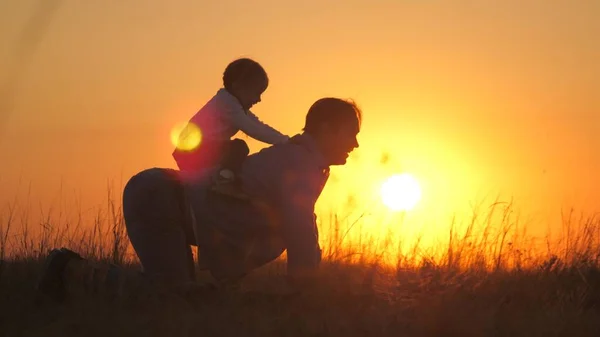 Тато грає зі своєю донькою в парку на траві під час заходу сонця. Батько їде за дитиною на спині, сидить осідлавши батька, радіє. Тато і маленька дитина бавляться надворі. Щаслива родина — стокове фото