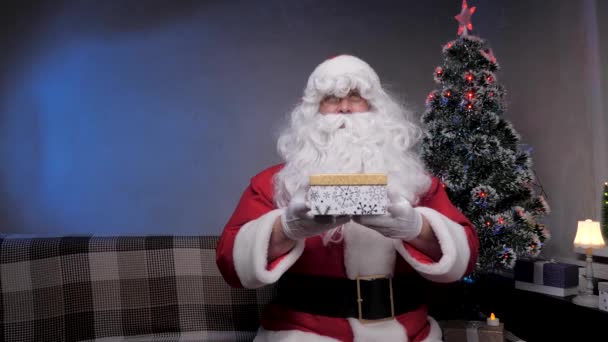 Санта Клаус сидить на дивані, тримаючи подарунок і кидаючи коробку спереду. Санта Клаус дарує подарунок аудиторії. Відпустка та святкування. Зимові сімейні канікули. Веселого Різдва. — стокове відео